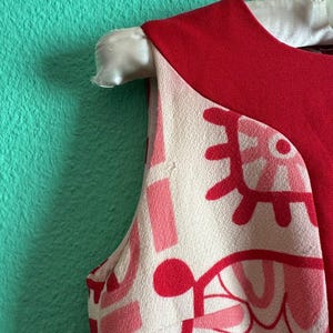 May include: A sleeveless dress featuring a solid red upper section and a cream-colored lower section with a bold, abstract graphic print in shades of pink and red. The garment has a round neckline and is displayed on a white hanger.