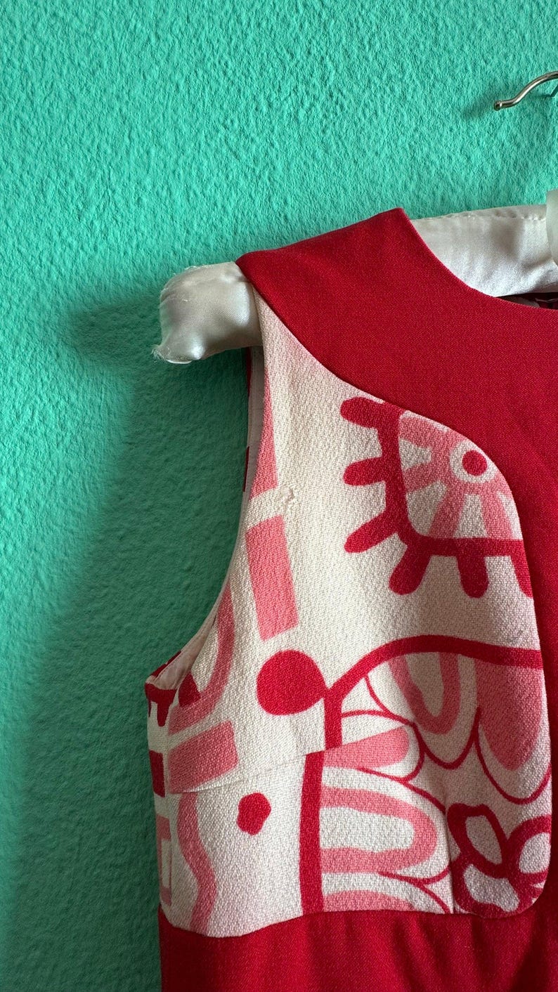 May include: A sleeveless dress featuring a solid red upper section and a cream-colored lower section with a bold, abstract graphic print in shades of pink and red. The garment has a round neckline and is displayed on a white hanger.