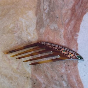 Può includere: Un pettine per capelli in tartaruga marrone dal design affusolato e appuntito. Il bordo superiore è decorato con una fila di piccole pietre trasparenti, motivi floreali arancioni con foglie verdi e intarsi in metallo argentato lungo il bordo.