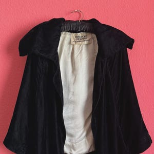 May include: A black velvet cape with a wide collar and an open front design. The interior is lined with white fabric and features a label that reads Bergdorf Goodman Fifth Avenue at 58th Street. The garment is displayed on a metal hanger.