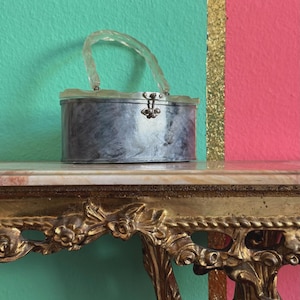 Könnte beinhalten: Eine silberfarbene Handtasche im Vintage-Stil mit transparentem Griff und Schnallenverschluss. Die Tasche steht auf einem Tisch mit Marmorplatte und goldenen Details. Der Hintergrund ist eine türkis- und rosafarbene Wand mit einem Goldstreifen.