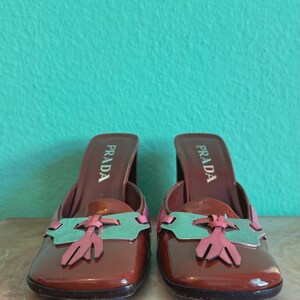 May include: A pair of brown leather Prada mules with a square toe and a block heel. The shoes feature a decorative design in pink and green on the vamp. The Prada logo is visible on the insole.