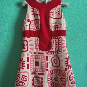 May include: A sleeveless dress featuring a cream-colored base with a repeating pattern of red and pink abstract geometric shapes. The garment includes a solid red panel across the chest and upper bodice, with a round neckline and a flared skirt.