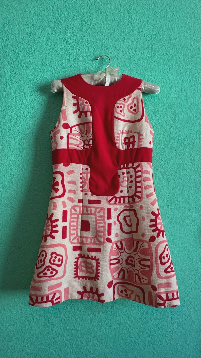 May include: A sleeveless dress featuring a cream-colored base with a repeating pattern of red and pink abstract geometric shapes. The garment includes a solid red panel across the chest and upper bodice, with a round neckline and a flared skirt.