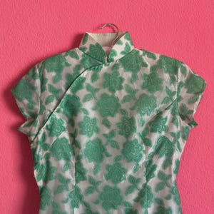 May include: A white and green floral patterned dress with short sleeves and a mandarin collar. The dress is hanging on a silver hanger against a pink background. The floral pattern is a repeating design.