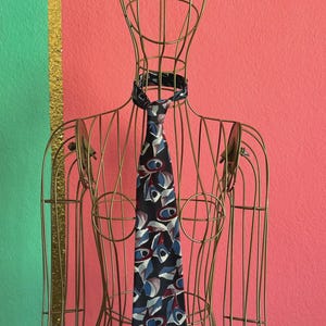 Pode incluir: Uma gravata com fundo azul-marinho escuro e um padrão abstrato repetitivo de formas geométricas brancas, azul-claras e bordô. A gravata é exibida num busto de manequim de arame dourado com um design de estrutura aberta.