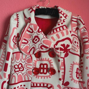 May include: A double-breasted coat featuring a cream base with a repeating graphic pattern in shades of red and pink. The coat has a wide collar, long sleeves, and four large, round, glossy red buttons arranged in two vertical rows on the front.
