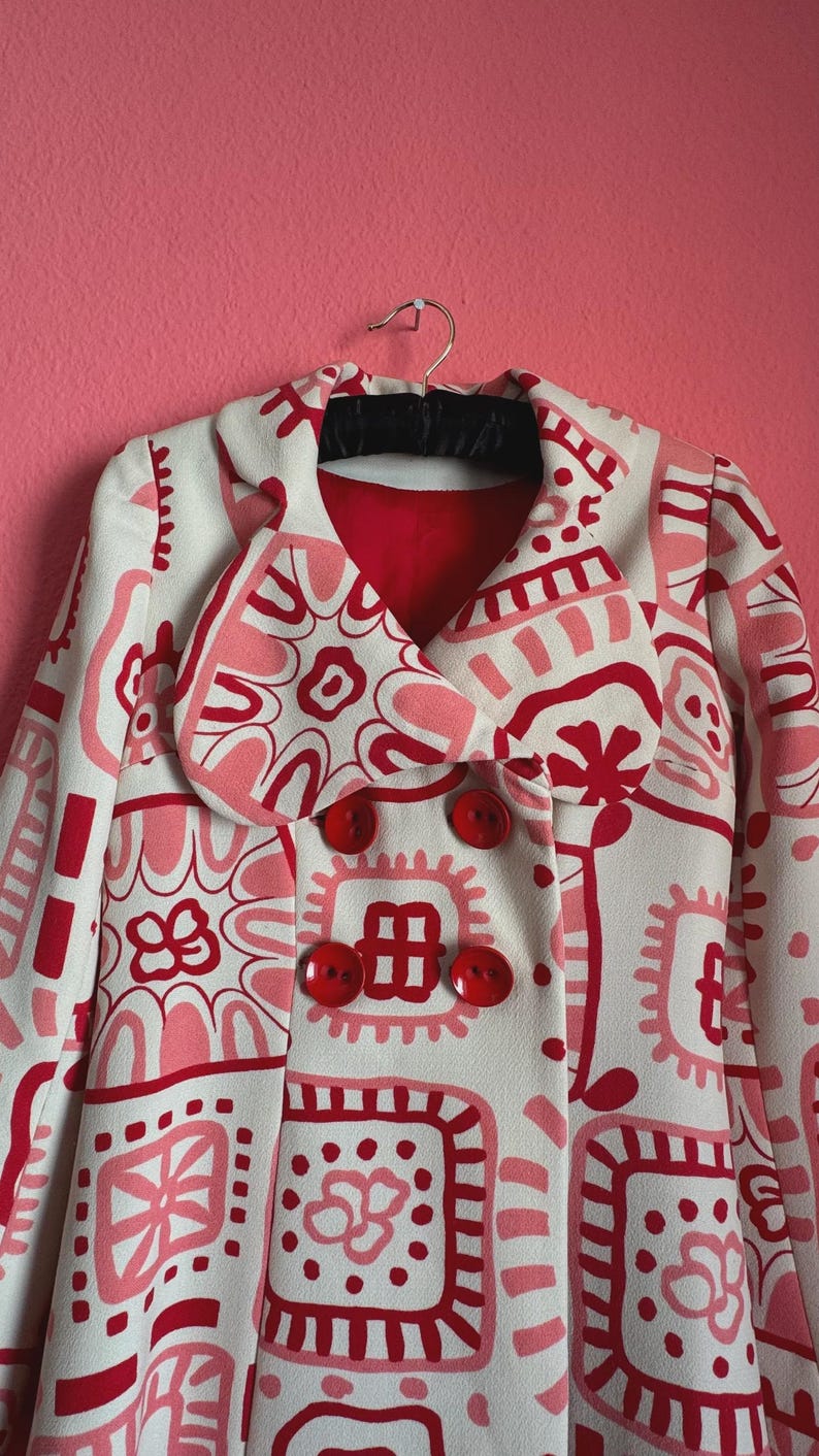 May include: A double-breasted coat featuring a cream base with a repeating graphic pattern in shades of red and pink. The coat has a wide collar, long sleeves, and four large, round, glossy red buttons arranged in two vertical rows on the front.