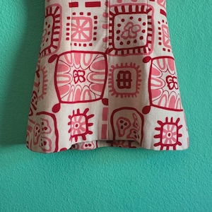May include: A sleeveless dress featuring a cream-colored base with a repeating pattern of red and pink geometric and floral squares. A solid red horizontal band sits across the chest area, and the garment has a straight, structured silhouette.