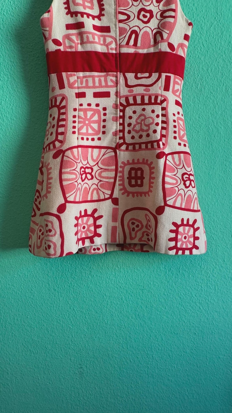 May include: A sleeveless dress featuring a cream-colored base with a repeating pattern of red and pink geometric and floral squares. A solid red horizontal band sits across the chest area, and the garment has a straight, structured silhouette.