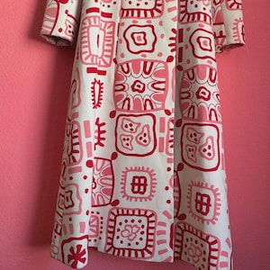 May include: A long-sleeved coat featuring a cream-colored base with a repeating pattern of square shapes in shades of pink and dark red. The coat has a wide collar and two large, round, dark red buttons positioned at the upper front closure.