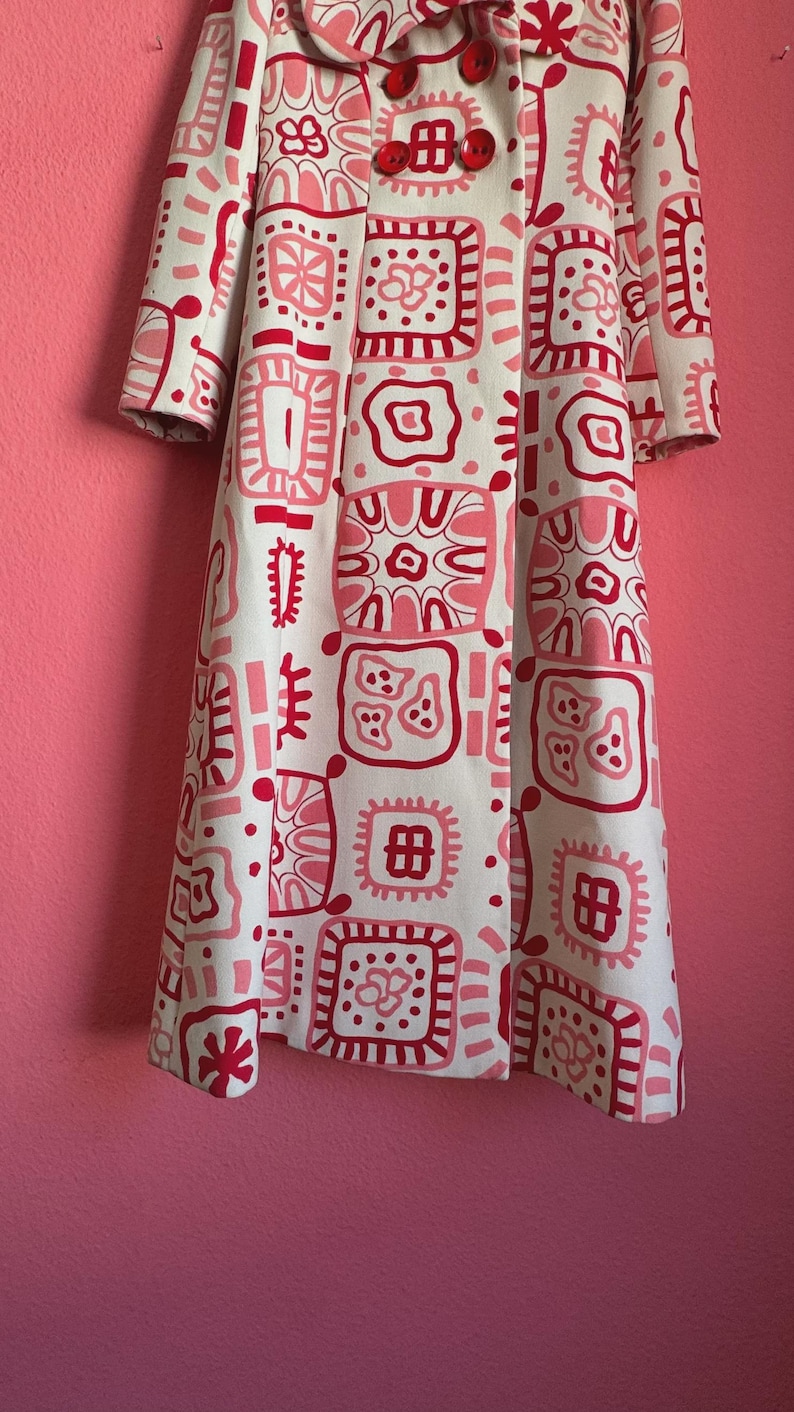 May include: A long-sleeved coat featuring a cream-colored base with a repeating pattern of square shapes in shades of pink and dark red. The coat has a wide collar and two large, round, dark red buttons positioned at the upper front closure.