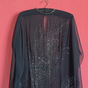 Könnte beinhalten: Ein schwarzes Langarmkleid aus transparentem Stoff mit hohem Ausschnitt und Schlüsselloch-Ausschnitt. Das Kleidungsstück ist mit einem dichten Muster aus kleinen, reflektierenden silbernen Strasssteinen verziert, die sich in der Mitte konzentrieren.