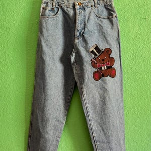 Può includere: Jeans di denim blu chiaro con una toppa a forma di orso con cilindro e papillon. I jeans sono appesi a una gruccia di velluto nero su sfondo verde. La toppa dell'orso è marrone, rossa e nera.