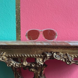 Può includere: Occhiali da sole in stile vintage con montatura bianca e lenti colorate appoggiati su una mensola in marmo. La mensola è sostenuta da una staffa dorata ornata. Lo sfondo presenta un design a blocchi di colore turchese, rosa e oro.