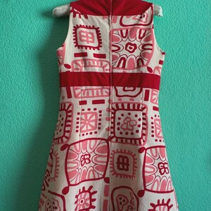 May include: A sleeveless dress in cream, featuring a repeating pattern of red and pink geometric shapes and floral motifs. The garment includes a solid red horizontal band across the waist and a vertical zipper closure along the center back.