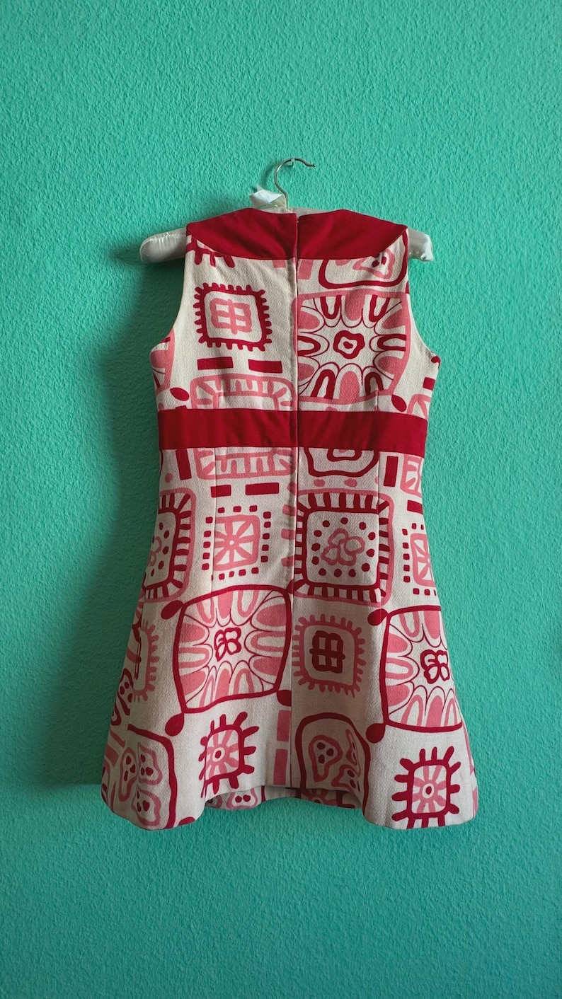 May include: A sleeveless dress in cream, featuring a repeating pattern of red and pink geometric shapes and floral motifs. The garment includes a solid red horizontal band across the waist and a vertical zipper closure along the center back.