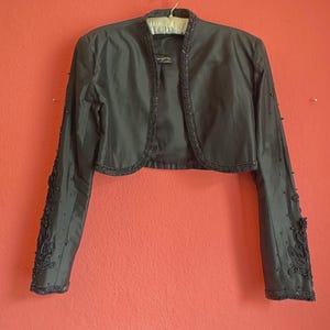 Può includere: Bolero grigio scuro con maniche lunghe, impreziosito da perline e ricami. La giacca è appesa a un gancio su uno sfondo corallo. Il bolero ha uno scollo tondo e una lunghezza corta.