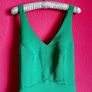 Può includere: Un abito verde smeraldo senza maniche con scollo a V è appeso a una gruccia imbottita in raso contro un muro rosa acceso. L'abito presenta un corpetto aderente e una silhouette semplice ed elegante, ideale per occasioni formali.