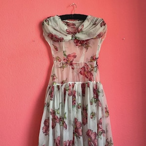 May include: A sleeveless dress featuring a sheer white overlay with a pink and green floral print. The bodice has a draped, folded neckline, and the skirt is gathered at the waist, falling to a mid-calf length. The dress is hung on a black hanger.