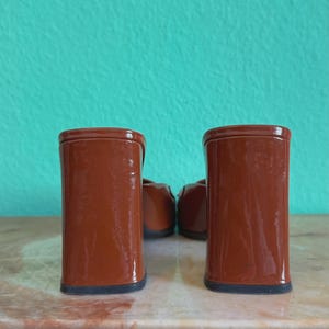 May include: A pair of brown heeled shoes with a block heel. The shoes are a glossy, reddish-brown color and have a black sole. The background is a teal wall.