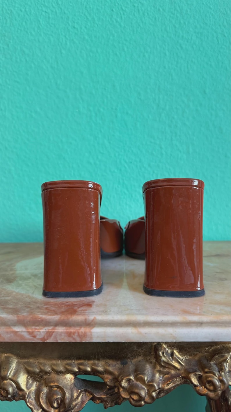 May include: A pair of brown heeled shoes with a block heel. The shoes are a glossy, reddish-brown color and have a black sole. The background is a teal wall.