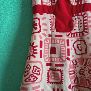 May include: A sleeveless dress featuring a cream-colored base with a repeating pattern of red and pink geometric shapes and abstract motifs. The garment includes a solid red panel across the chest and a round neckline, showcasing a textured fabric.