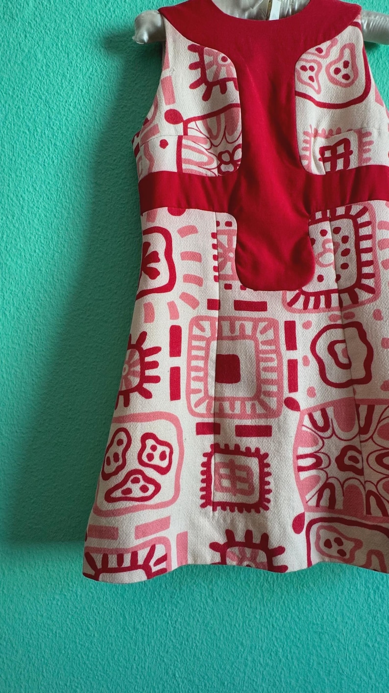 May include: A sleeveless dress featuring a cream-colored base with a repeating pattern of red and pink geometric shapes and abstract motifs. The garment includes a solid red panel across the chest and a round neckline, showcasing a textured fabric.