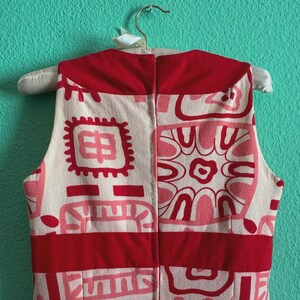 May include: A sleeveless dress featuring a cream base with a repeating geometric pattern in shades of red and pink. The garment includes a solid red horizontal band at the waist and a solid red collar section, with a vertical zipper closure at the back.