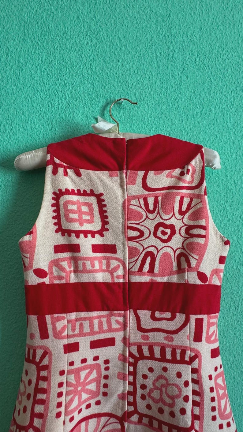 May include: A sleeveless dress featuring a cream base with a repeating geometric pattern in shades of red and pink. The garment includes a solid red horizontal band at the waist and a solid red collar section, with a vertical zipper closure at the back.
