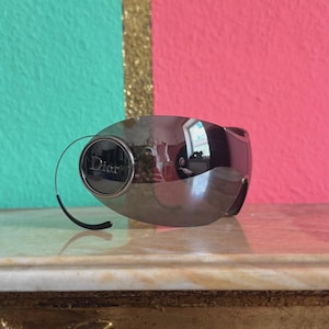 May include: Silver-toned Dior sunglasses with a curved, wraparound design. The sunglasses have a dark lens and a circular accent on the side with the Dior logo. The background is a mix of teal, gold, and pink.