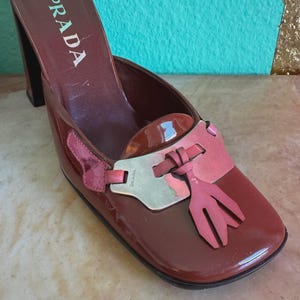 May include: A brown Prada heeled mule with a square toe. The shoe features a pink accent with a tassel design and a silver-tone detail. The Prada logo is visible on the side of the shoe.