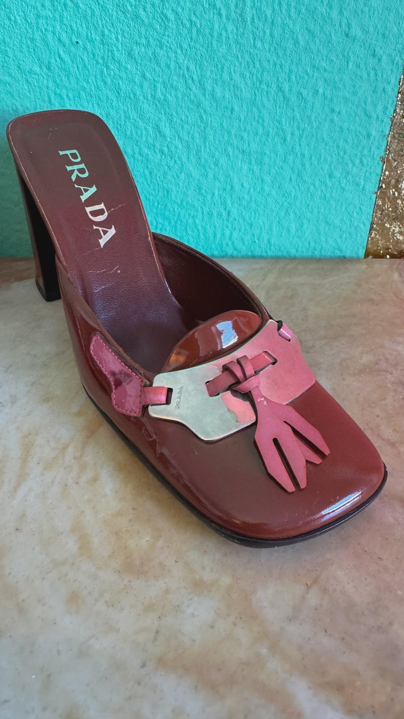 May include: A brown Prada heeled mule with a square toe. The shoe features a pink accent with a tassel design and a silver-tone detail. The Prada logo is visible on the side of the shoe.