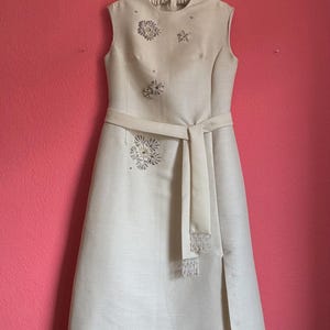 May include: A sleeveless, floor-length cream-colored dress featuring a high neckline, a matching tie-waist belt, and a side slit. The bodice is adorned with scattered, circular silver-toned beaded floral embellishments and small rhinestones.