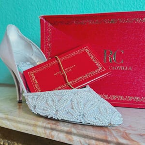 May include: A white beaded shoe with a high heel, a red box, and a red card with gold lettering. The shoe is decorated with small white beads. The red box has the letters "RC" and the name "CAOVILLA" in gold.