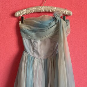 May include: A vintage strapless gown in shades of blue and white hangs on a satin-covered hanger against a pink background. The dress features a draped bodice and a flowing, layered skirt, creating an elegant silhouette. The fabric appears to be a delicate chiffon or tulle.