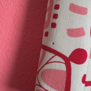 May include: A textured fabric featuring a repeating abstract pattern in shades of light pink, dark pink, and red on a white background. The material displays a small, singular dark pinhole imperfection near the center of the visible surface area.
