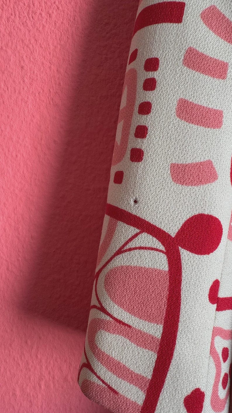 May include: A textured fabric featuring a repeating abstract pattern in shades of light pink, dark pink, and red on a white background. The material displays a small, singular dark pinhole imperfection near the center of the visible surface area.