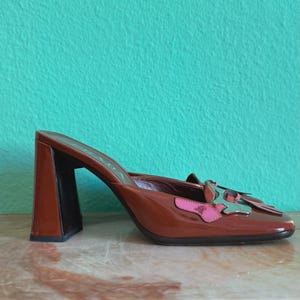 May include: Brown leather heeled mules with a square toe and a block heel. The shoe features a decorative accent with pink and silver details. The background is a solid teal color.