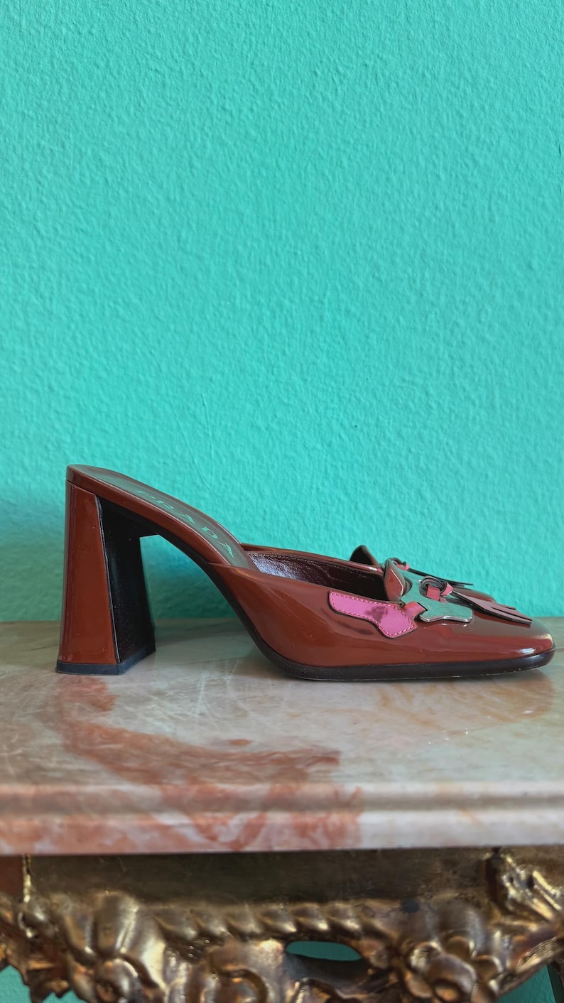 May include: Brown leather heeled mules with a square toe and a block heel. The shoe features a decorative accent with pink and silver details. The background is a solid teal color.