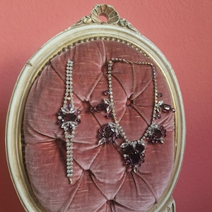 Può includere: Un set di gioielli vintage con un braccialetto e una collana con accenti di cristallo viola e trasparente. I gioielli sono esposti su una sedia di velluto rosa con una cornice decorativa e ornata. La sedia è posta su uno sfondo rosso.