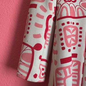 May include: A long-sleeved coat featuring an off-white base color with a bold, abstract graphic pattern in shades of bright red and soft pink. The design consists of various geometric shapes, curved lines, and floral-inspired motifs.