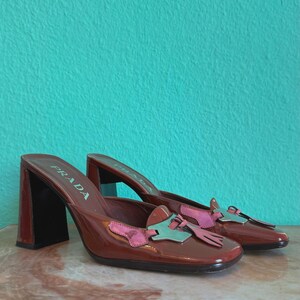 May include: A pair of brown leather Prada heeled mules. The shoes have a square toe and a block heel. The vamp features a pink and teal bow detail. The shoes are displayed on a marble surface against a teal background.
