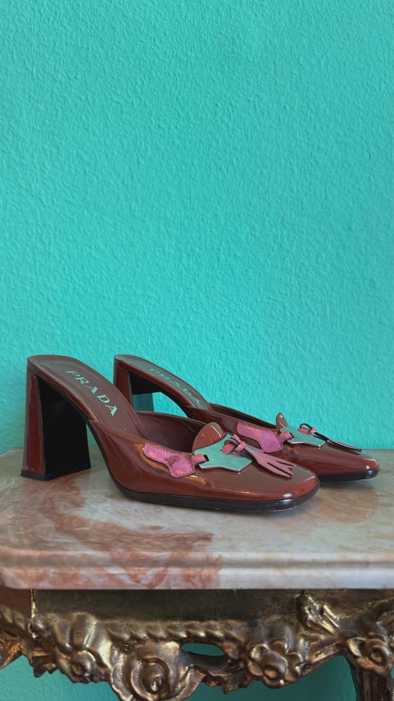 May include: A pair of brown leather Prada heeled mules. The shoes have a square toe and a block heel. The vamp features a pink and teal bow detail. The shoes are displayed on a marble surface against a teal background.