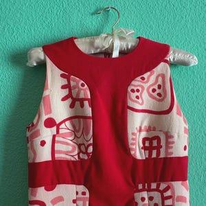 May include: A sleeveless dress featuring a cream-colored base with a repeating pattern of pink and red abstract shapes. The garment includes a solid red panel extending from the round neckline down the center and a horizontal red band at the waist.