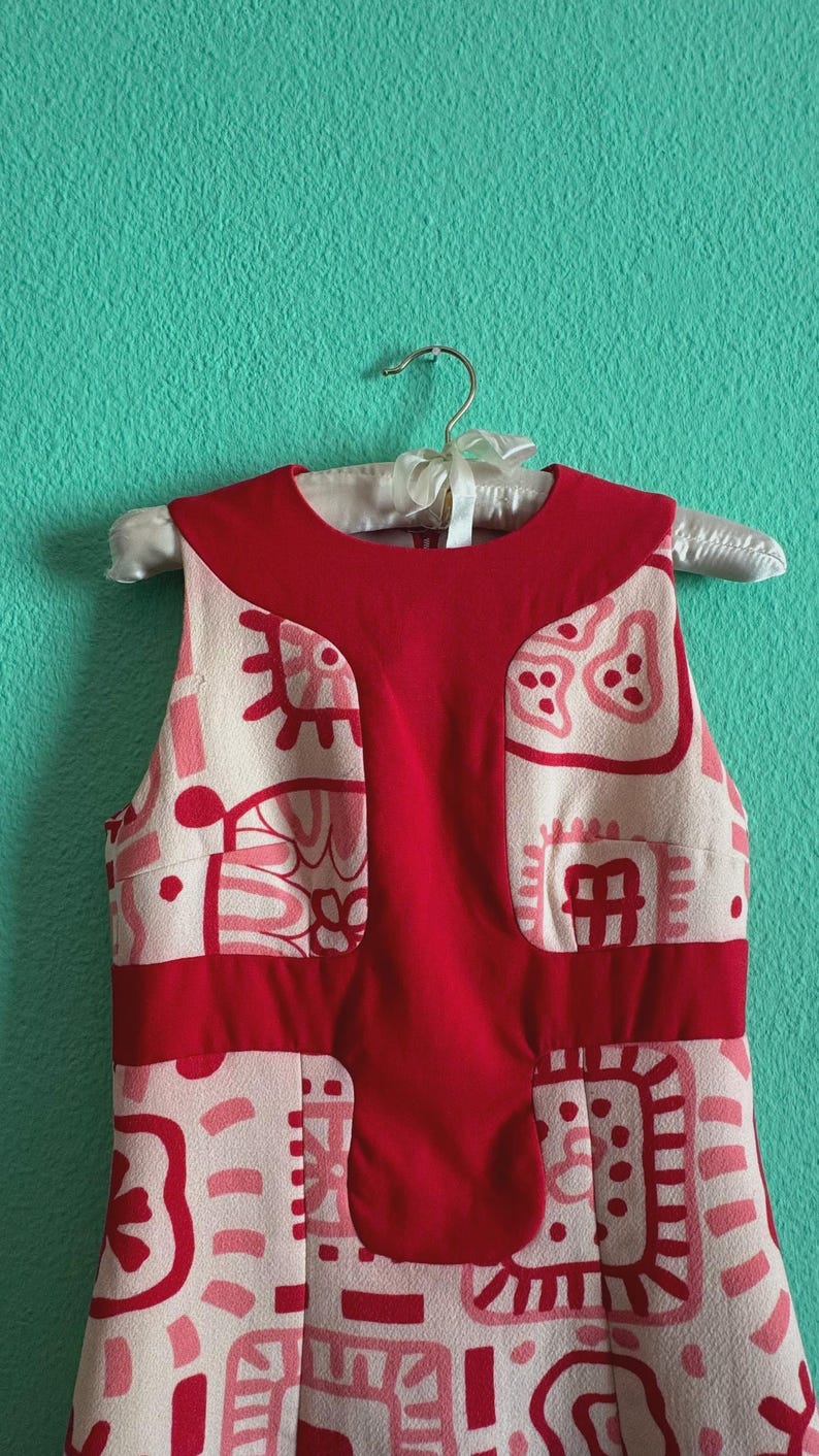 May include: A sleeveless dress featuring a cream-colored base with a repeating pattern of pink and red abstract shapes. The garment includes a solid red panel extending from the round neckline down the center and a horizontal red band at the waist.