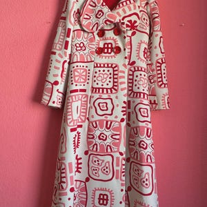 May include: A long-length coat in off-white featuring a repeating pattern of squares and abstract shapes in shades of pink and red. The coat has a wide collar, long sleeves, and three large, round, bright red buttons down the front center.