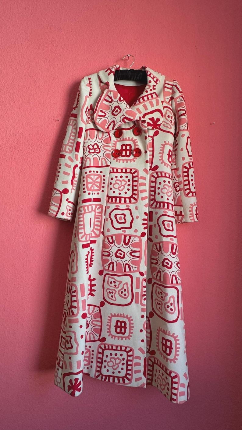 May include: A long-length coat in off-white featuring a repeating pattern of squares and abstract shapes in shades of pink and red. The coat has a wide collar, long sleeves, and three large, round, bright red buttons down the front center.