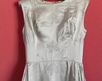 May include: Ivory-colored vintage dress with floral embroidery. The sleeveless dress hangs on a hanger against a pink background. The dress features a fitted bodice and a flared skirt, with embroidered floral patterns throughout.