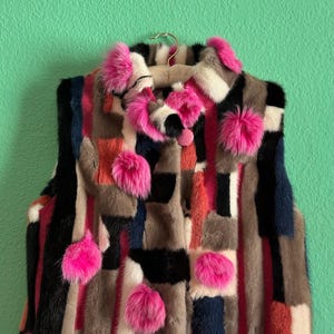 Puede incluir: Un chaleco de piel de retazos colorido con una variedad de colores que incluyen negro, rosa, rojo y gris. El chaleco está adornado con pompones rosas brillantes y cuelga de una percha de madera sobre un fondo verde.